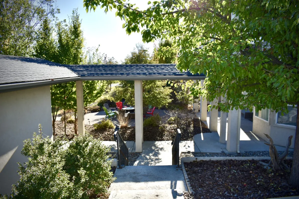A colorful, inviting patio framed by lush greenery, with bright chairs arranged for relaxation. The pathway leads toward the patio, seamlessly connecting indoor and outdoor spaces, and the surrounding foliage creates a private, resort-like setting. The patio is ideal for enjoying views, morning coffee, or a celebratory barbecue after a day on the golf course.