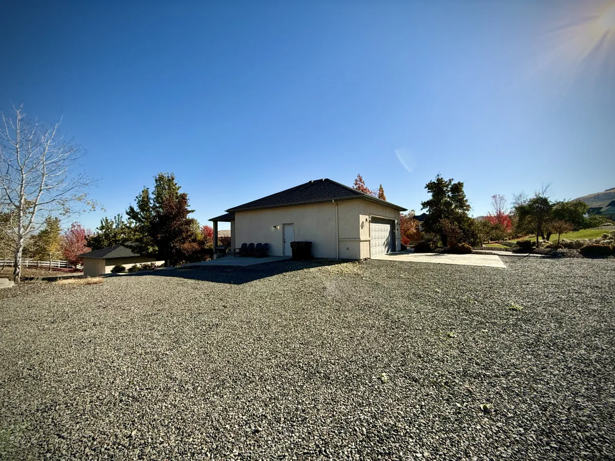 An exterior shot featuring the house set against a backdrop of trees, with a gravel driveway and calm natural surroundings. The landscape hints at the foothill location and the serene, private environment awaiting guests.