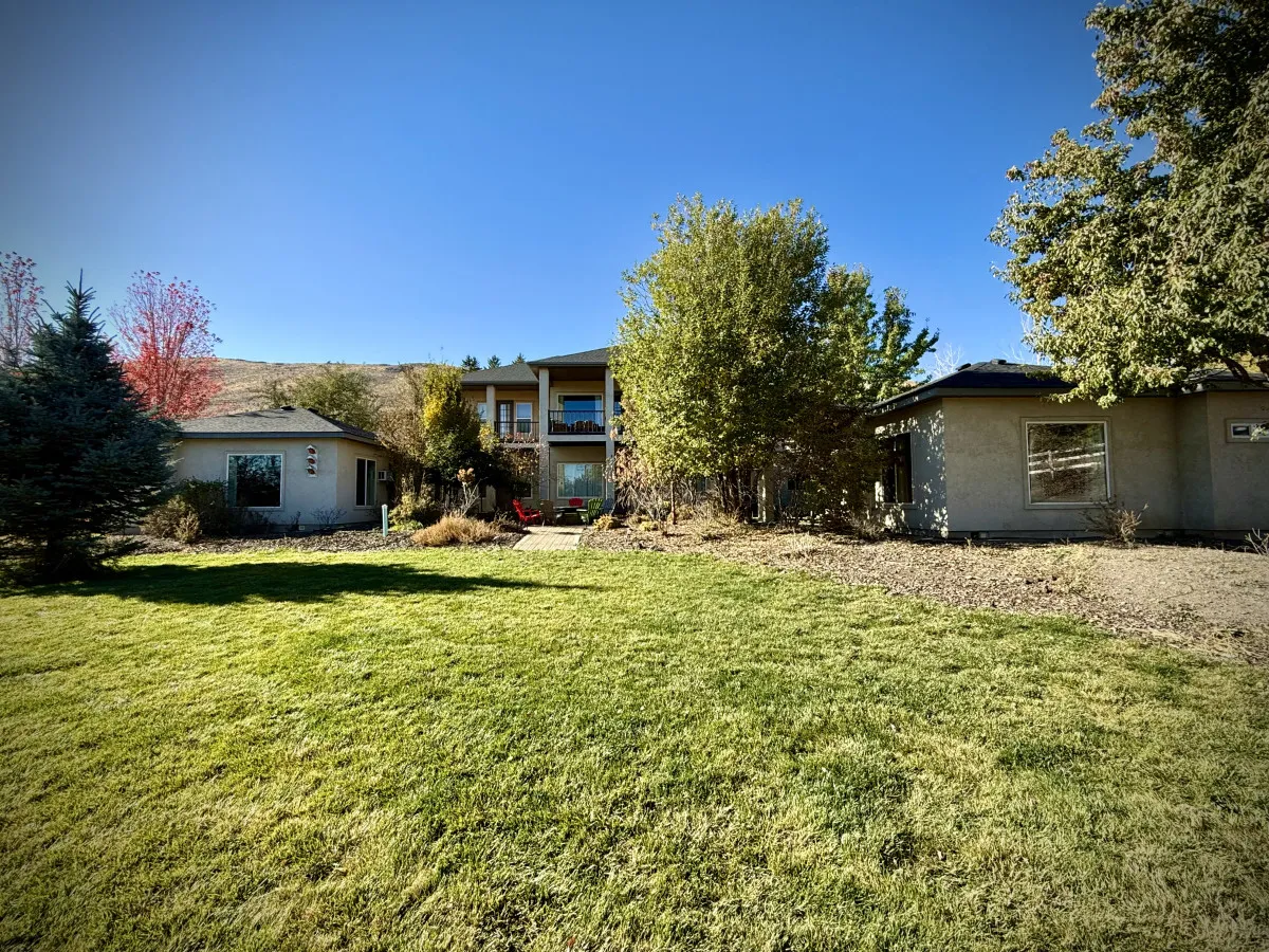 The image showcases both the main house and a secondary structure set amidst well-kept landscaping and mature trees. The multiple buildings clearly suggest an estate-like property, ideal for accommodating larger groups, retreats, or exclusive bookings. The open, tranquil yard hints at ample space for outdoor activities and relaxation after a day on the links.