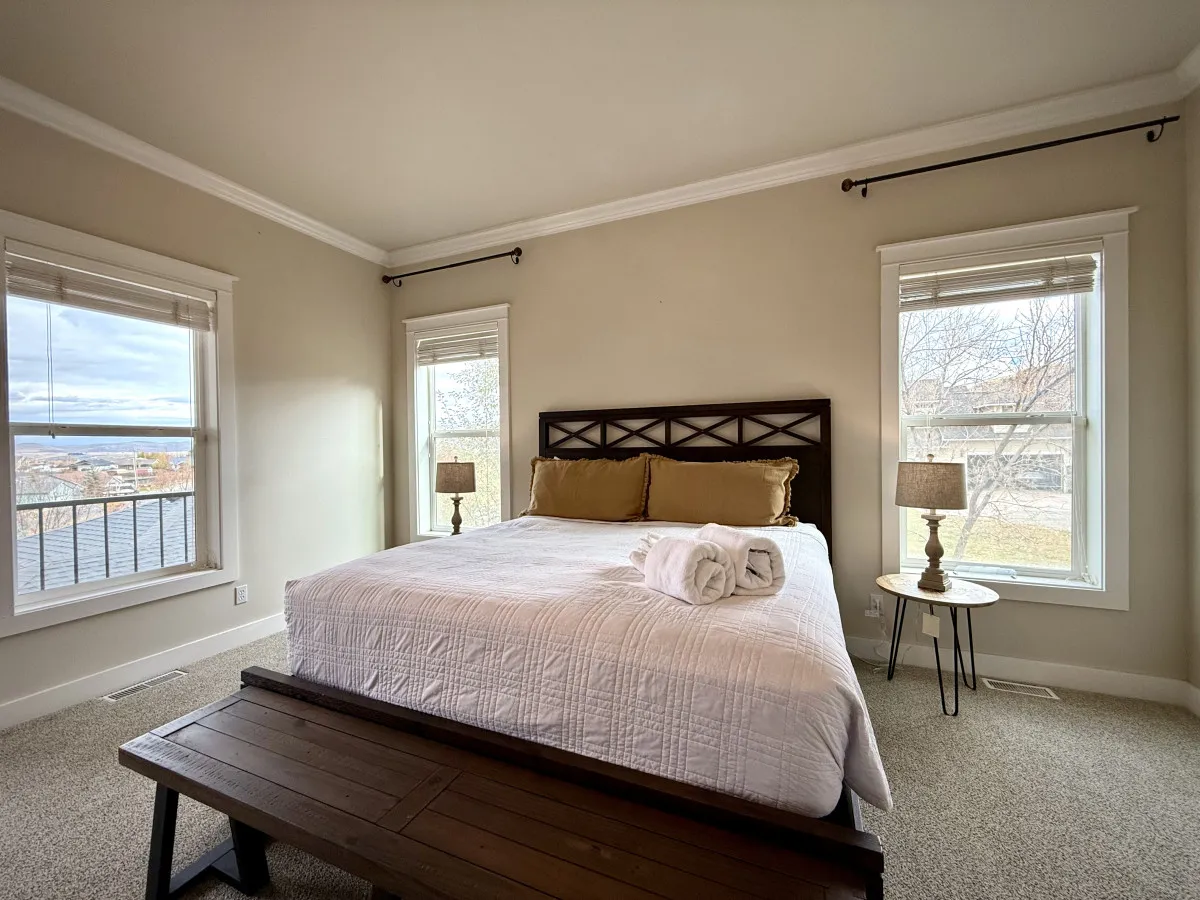 A peaceful bedroom with a plush bed adorned with pillows, large windows letting in scenic natural light, and nightstands with matching lamps, all set in a tranquil color palette.