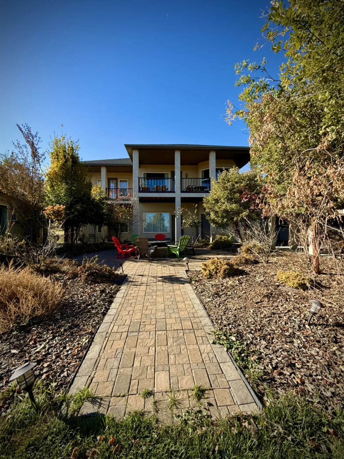 Two colorful chairs, one red and one green, are positioned on a pathway leading toward the home, with the house and surrounding garden visible. The chairs are set near an outdoor fire pit, suggesting a welcoming space for gathering after outdoor activities.