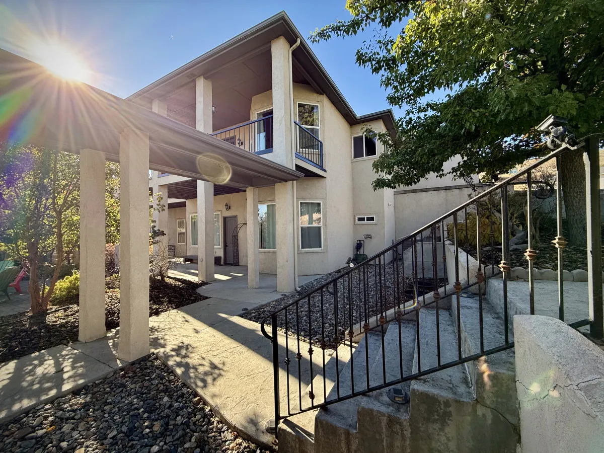 A wide view of the home's exterior featuring entrance steps, a balcony, and lush surrounding greenery. The scene suggests both accessibility and inviting outdoor spaces for gathering, with the balcony hinting at views perfect for sunset toasts or morning coffee.