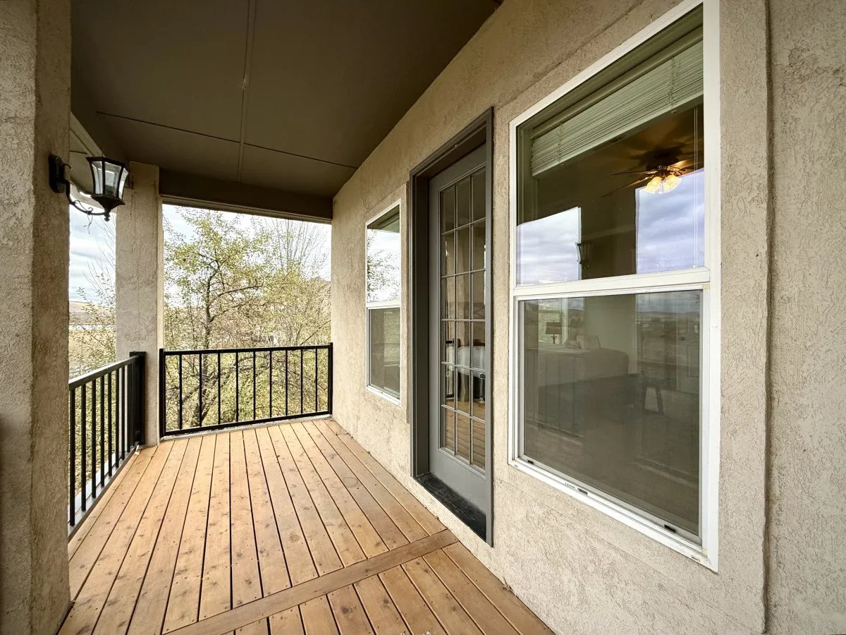 A tranquil exterior shot of a wooden deck bordered by a sleek black railing, with a glass-paned door leading inside and lush trees surrounding the space. The lighting hints at the calm of morning or dusk, suggesting an ideal spot for reflection or relaxation.