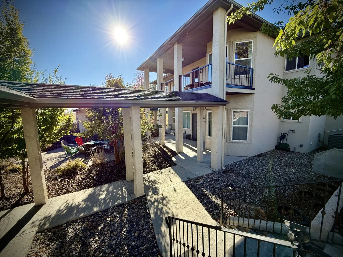 The image showcases the exterior of a spacious single-family home bathed in warm sunlight, with a charming porch supported by classic columns. Lush green foliage surrounds the house, and a comfortable outdoor seating area invites guests to relax and enjoy the scenic foothill views. The atmosphere is tranquil and welcoming, perfect for gatherings or quiet moments.