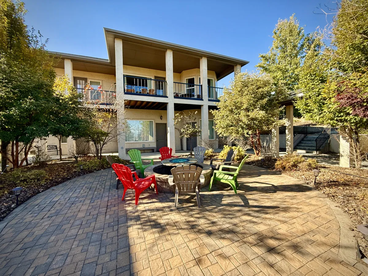 A vibrant patio scene with a fire pit surrounded by colorful chairs, an upper balcony visible above, and a lush green setting. The area is set up for gatherings, meals, and sunset views, embodying the outdoor relaxation and entertainment theme.