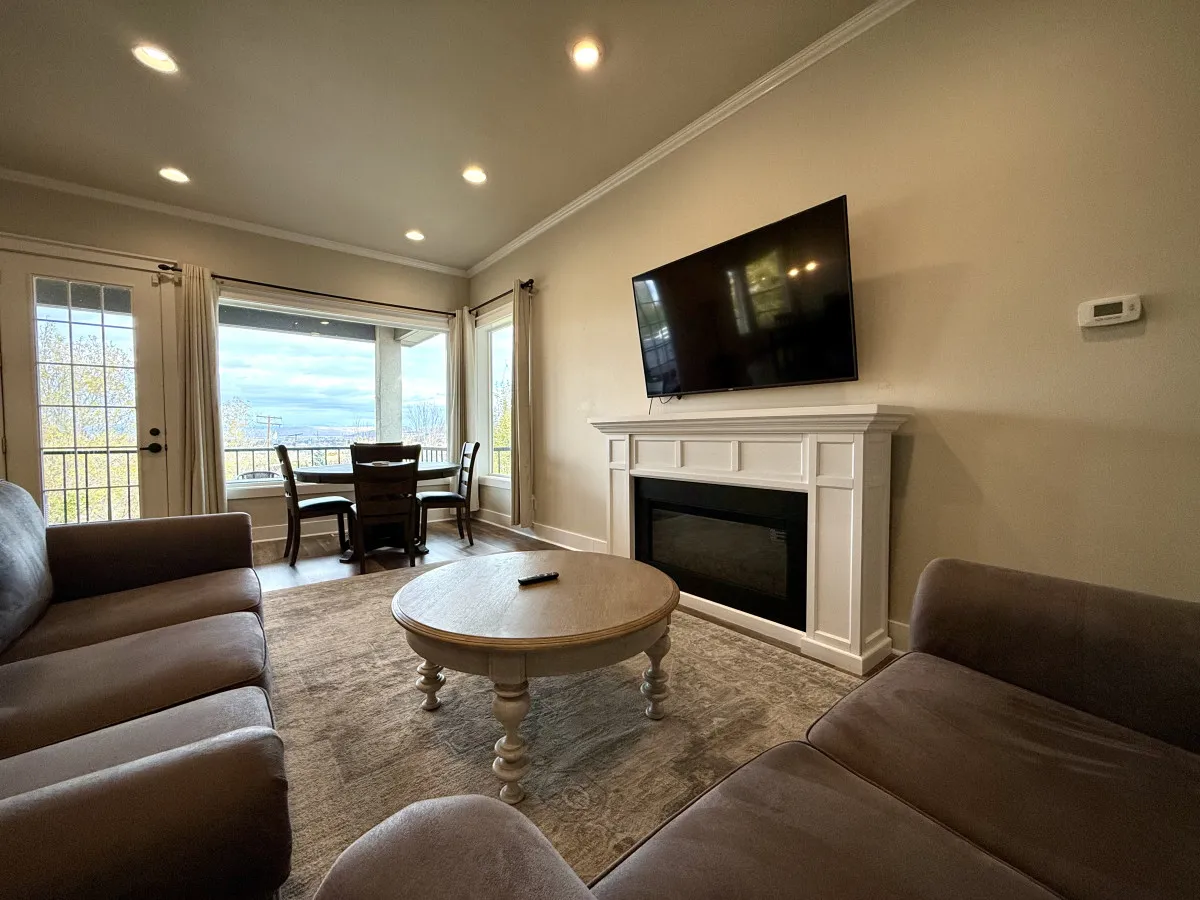 A cozy basement living area featuring a comfortable sofa, large TV, fireplace, and a dining area with views through the windows. The space is designed for family gatherings, movie nights, and casual dining, offering a warm and inviting atmosphere.