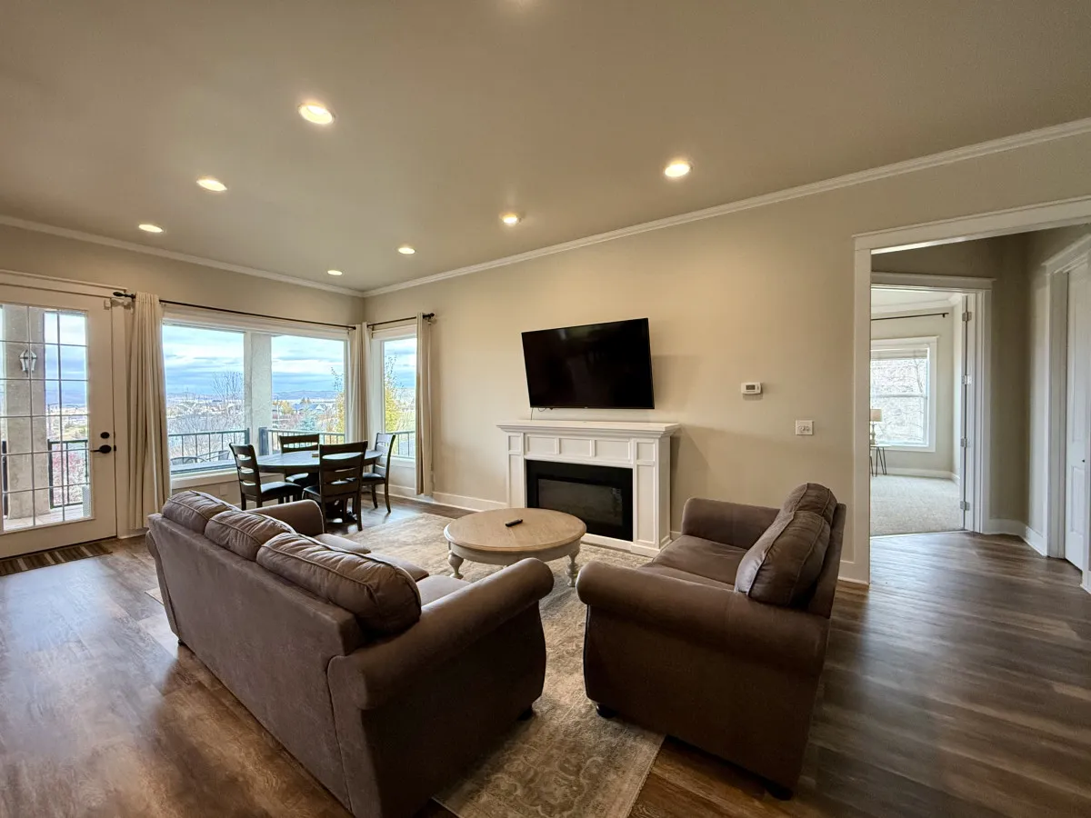 A cozy and inviting living space with large windows showcasing scenic views, a comfortable seating area, a modern fireplace, and a dining table. The room is bathed in natural light, enhancing its warmth and elegance, and providing a place for both relaxation and social gatherings.