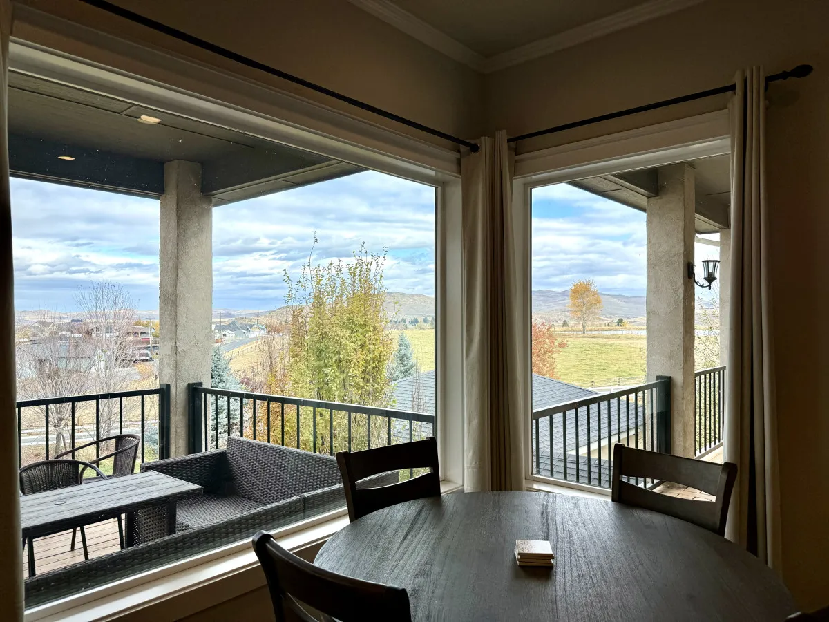 A dining area is featured with a table set beside large windows, offering scenic outdoor views and abundant natural light. Outdoor seating is visible beyond, reinforcing the sense of connection to nature and flexible gathering spaces.
